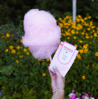 A hand holds a pink cotton candy stick with its paper tag attached, set against a blurred background of green foliage and yellow flowers. The tag reads 'PEARL cotton candy'.